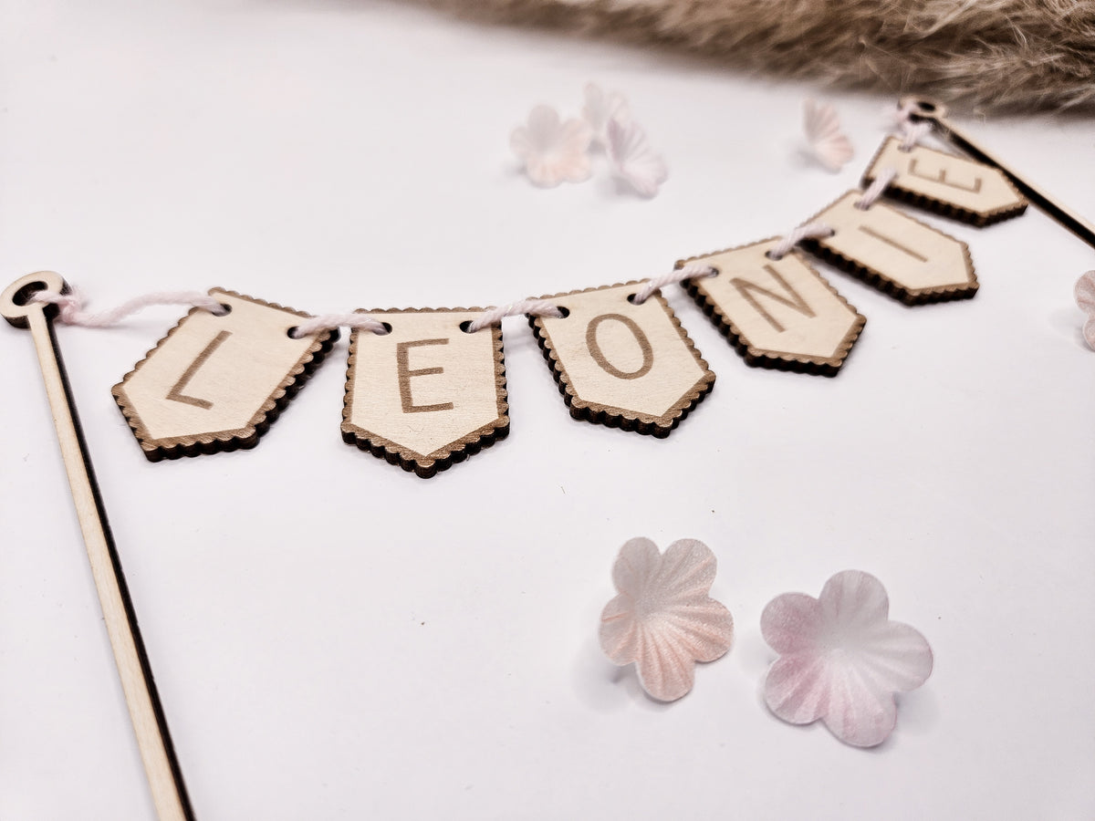 Personalisierter Cake Topper aus Holz mit dem Namen „Leonie“ als Wimpelgirlande für eine Torte zur Taufe, Geburt oder zum Geburtstag. Zwei filigrane Holzstäbe tragen eine Girlande mit gravierten Buchstaben auf naturfarbenen Holzfähnchen. Die schlichte Naked Cake Torte ist mit zwei niedlichen Hasenfiguren und zarten rosafarbenen Blüten dekoriert. Das zeitlose, skandinavische Design macht den personalisierten Tortenstecker zu einem besonderen Highlight jeder Feier.