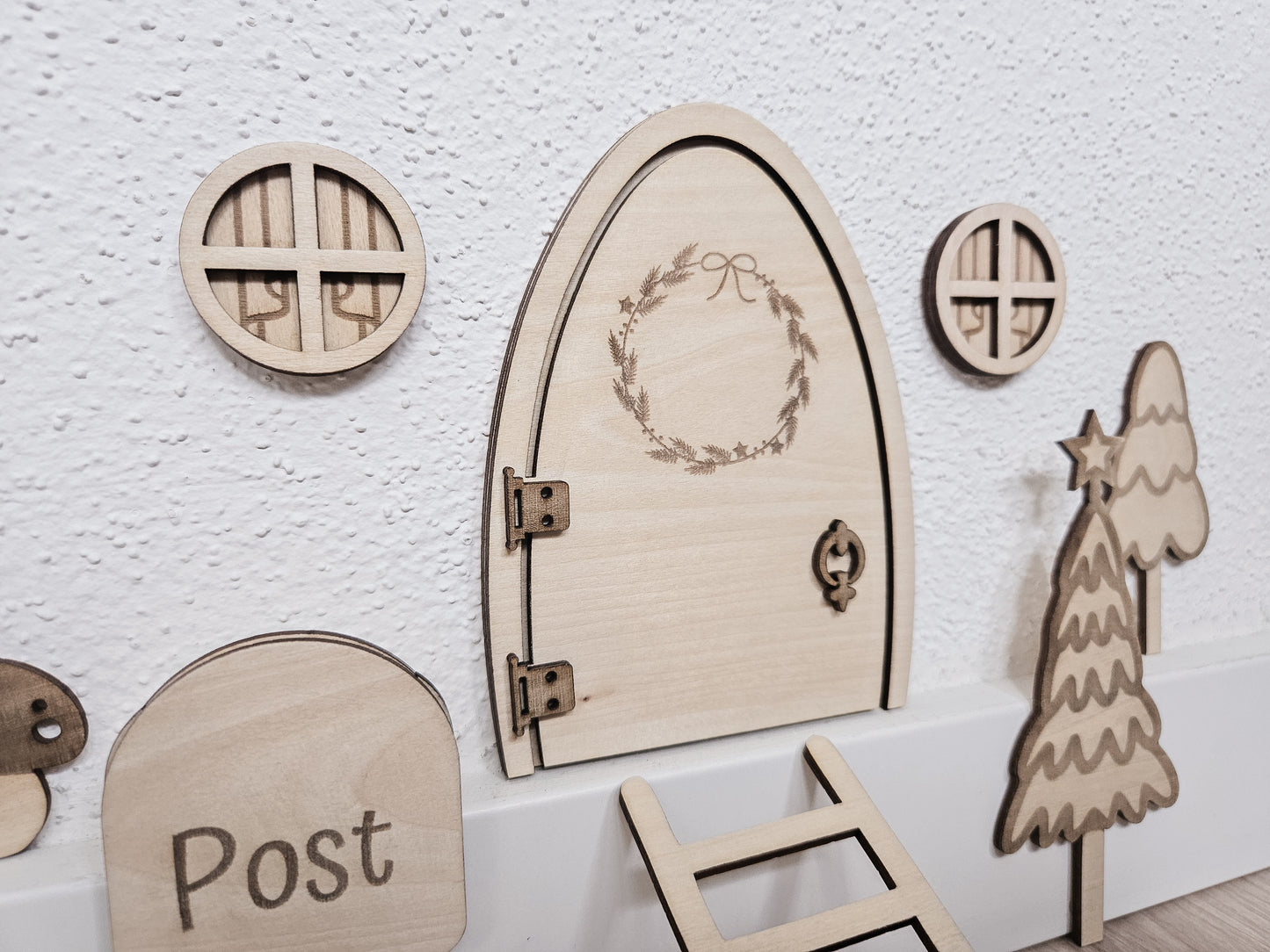 Personalisierbare Holz-Wichteltür mit Leiter, Briefkasten, Pilz und zwei Tannenbäumen an einer weißen Sockelleiste im Kinderzimmer, auf hellblauer Wand mit weißen Punkten montiert.