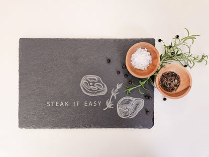 Schieferplatte mit der Aufschrift Steak it easy sowie zwei Steaks und Tymianzweig eingraviert. Zudem Dekoration.
