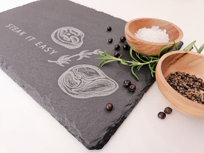 Schieferplatte mit der Aufschrift Steak it easy sowie zwei Steaks und Tymianzweig eingraviert. Zudem Dekoration.