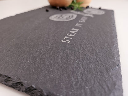 Schieferplatte mit der Aufschrift Steak it easy sowie zwei Steaks und Tymianzweig eingraviert. Zudem Dekoration.