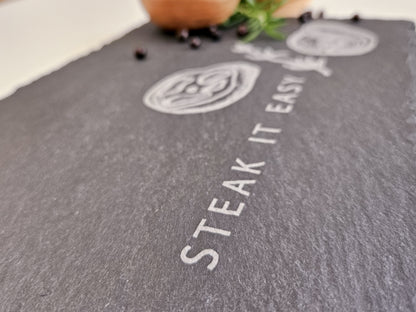 Schieferplatte mit der Aufschrift Steak it easy sowie zwei Steaks und Tymianzweig eingraviert. Zudem Dekoration.
