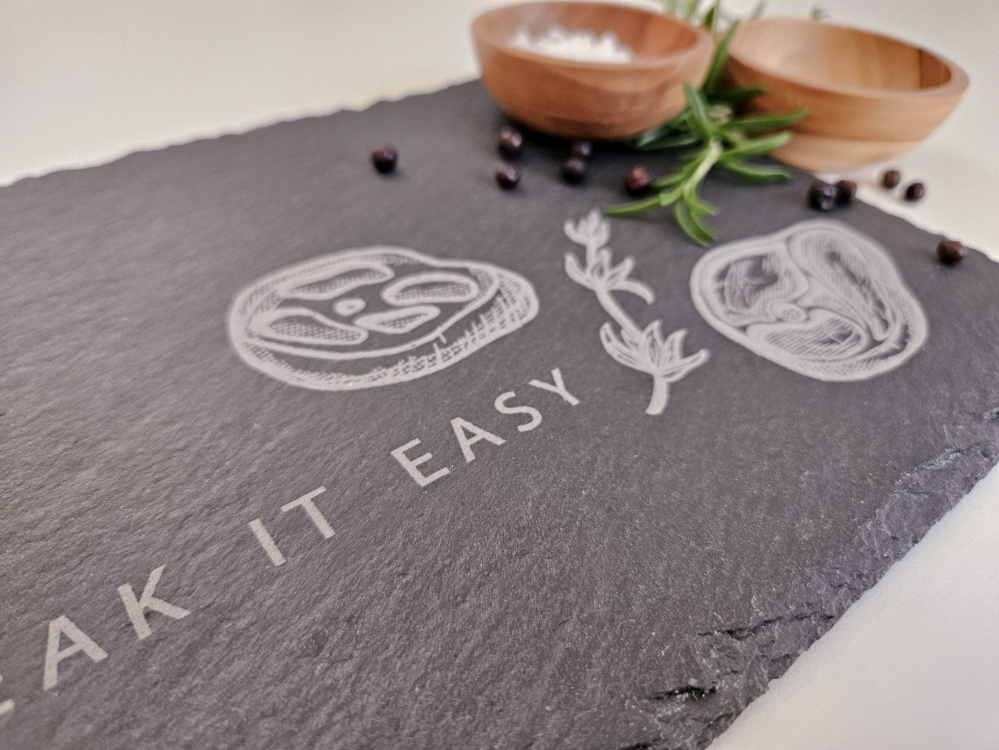 Schieferplatte mit der Aufschrift Steak it easy sowie zwei Steaks und Tymianzweig eingraviert. Zudem Dekoration.