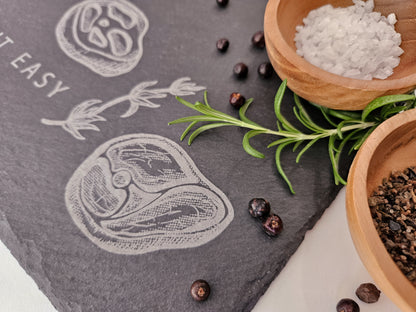 Schieferplatte mit der Aufschrift Steak it easy sowie zwei Steaks und Tymianzweig eingraviert. Zudem Dekoration.