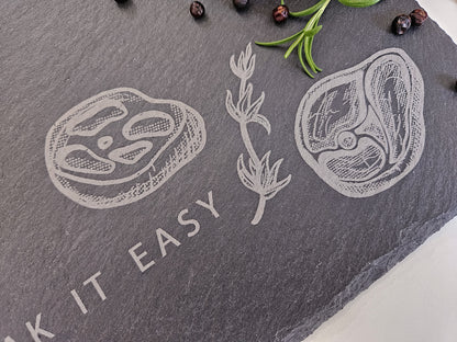 Schieferplatte mit der Aufschrift Steak it easy sowie zwei Steaks und Tymianzweig eingraviert. Zudem Dekoration.