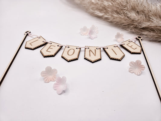 Personalisierter Cake Topper aus Holz mit dem Namen „Leonie“ als Wimpelgirlande für eine Torte zur Taufe, Geburt oder zum Geburtstag. Zwei filigrane Holzstäbe tragen eine Girlande mit gravierten Buchstaben auf naturfarbenen Holzfähnchen. Die schlichte Naked Cake Torte ist mit zwei niedlichen Hasenfiguren und zarten rosafarbenen Blüten dekoriert. Das zeitlose, skandinavische Design macht den personalisierten Tortenstecker zu einem besonderen Highlight jeder Feier.