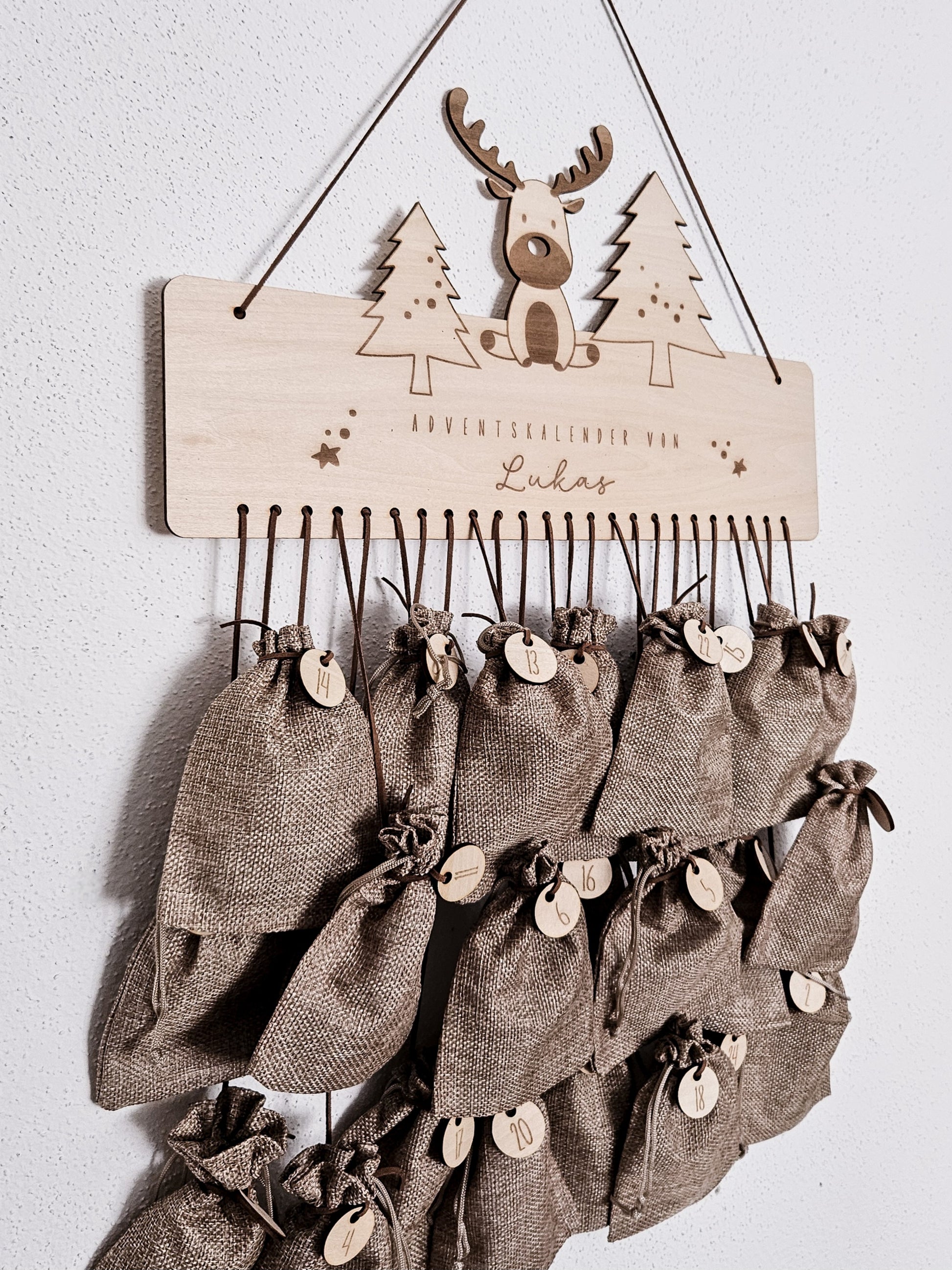 Personalisierter Holz-Adventskalender mit Rentier und Tannenbäumen, Gravur „Adventskalender von Lukas“. Mit 24 nummerierten Holzanhängern zum Aufhängen kleiner Geschenke. Nachhaltige Weihnachtsdekoration für Kinder und Familien.