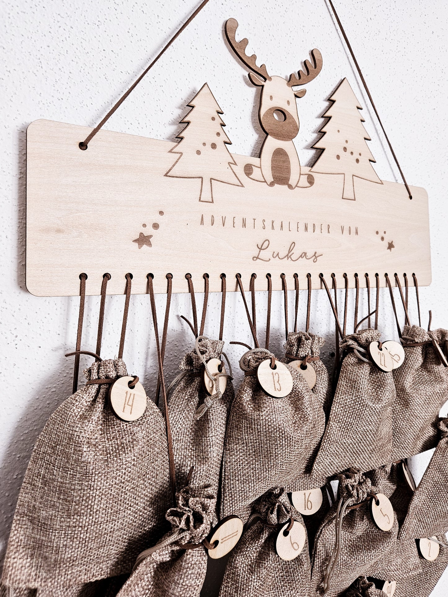 Personalisierter Holz-Adventskalender mit Rentier und Tannenbäumen, Gravur „Adventskalender von Lukas“. Mit 24 nummerierten Holzanhängern zum Aufhängen kleiner Geschenke. Nachhaltige Weihnachtsdekoration für Kinder und Familien.