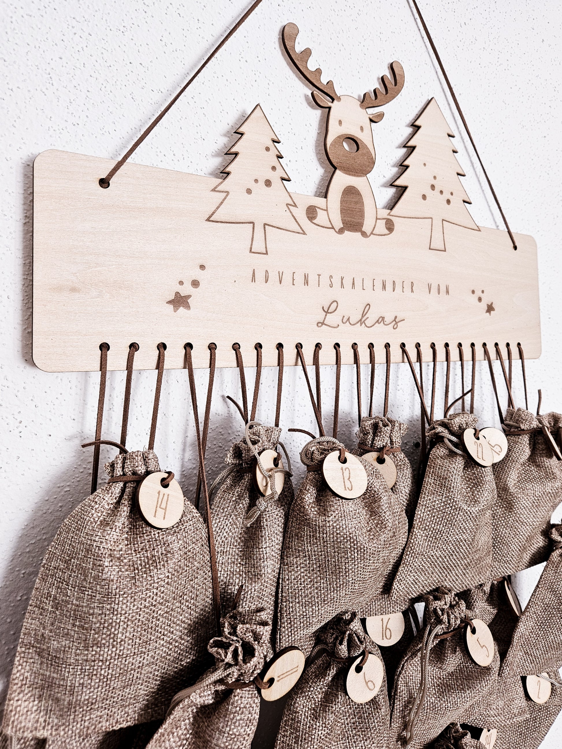Personalisierter Holz-Adventskalender mit Rentier und Tannenbäumen, Gravur „Adventskalender von Lukas“. Mit 24 nummerierten Holzanhängern zum Aufhängen kleiner Geschenke. Nachhaltige Weihnachtsdekoration für Kinder und Familien.