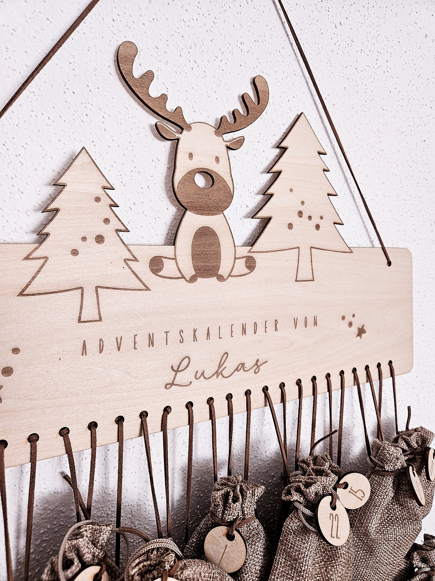 Personalisierter Holz-Adventskalender mit Rentier und Tannenbäumen, Gravur „Adventskalender von Lukas“. Mit 24 nummerierten Holzanhängern zum Aufhängen kleiner Geschenke. Nachhaltige Weihnachtsdekoration für Kinder und Familien.