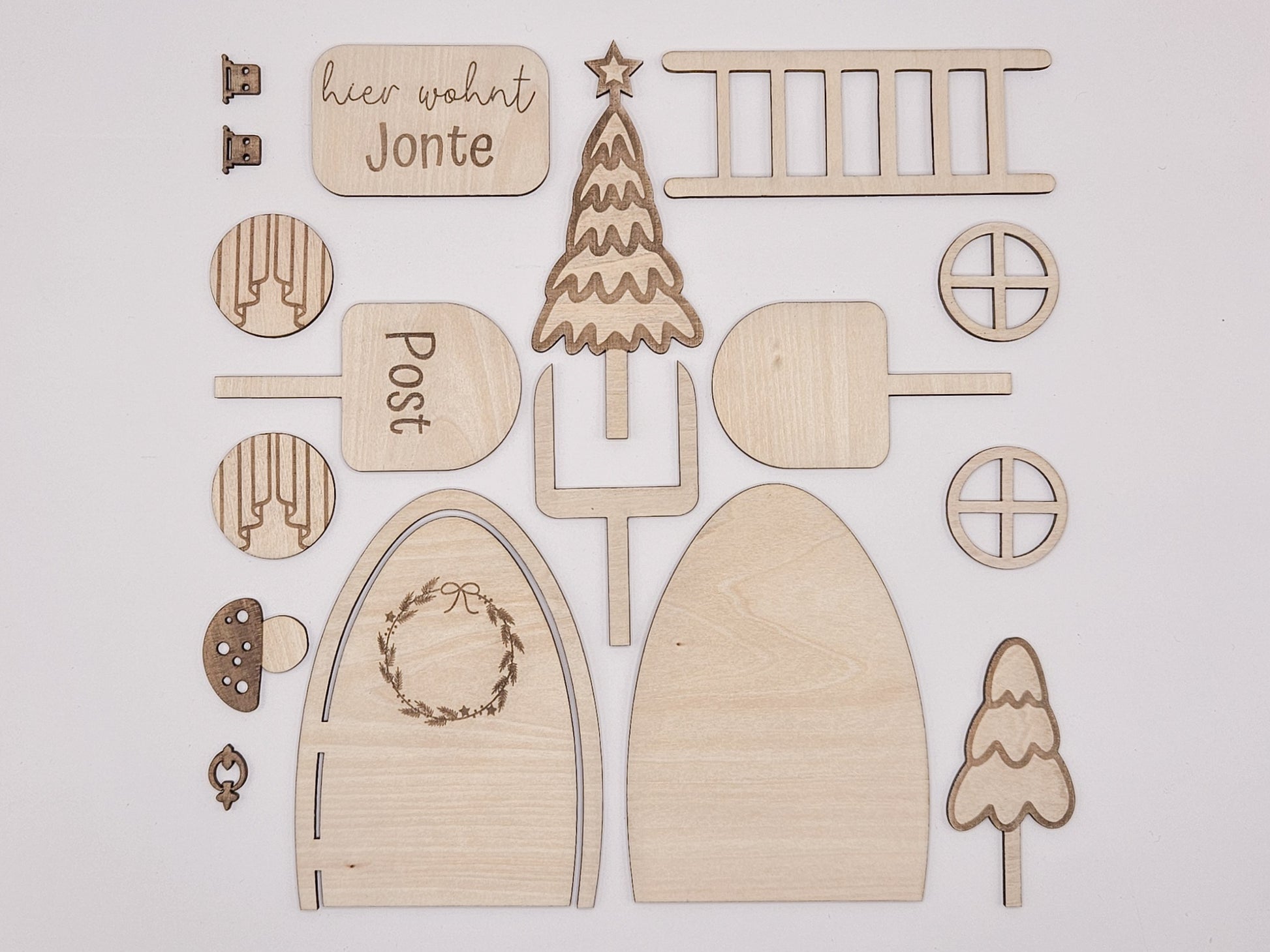 Personalisierbare Holz-Wichteltür mit Leiter, Briefkasten, Pilz und zwei Tannenbäumen an einer weißen Sockelleiste im Kinderzimmer, auf hellblauer Wand mit weißen Punkten montiert.