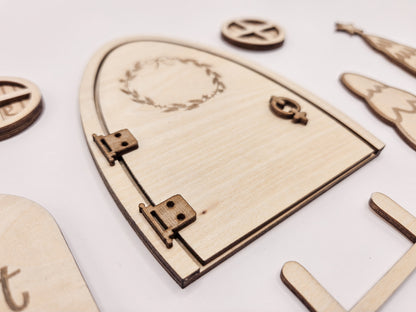 Personalisierbare Holz-Wichteltür mit Leiter, Briefkasten, Pilz und zwei Tannenbäumen an einer weißen Sockelleiste im Kinderzimmer, auf hellblauer Wand mit weißen Punkten montiert.