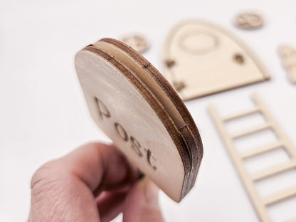 Personalisierbare Holz-Wichteltür mit Leiter, Briefkasten, Pilz und zwei Tannenbäumen an einer weißen Sockelleiste im Kinderzimmer, auf hellblauer Wand mit weißen Punkten montiert.