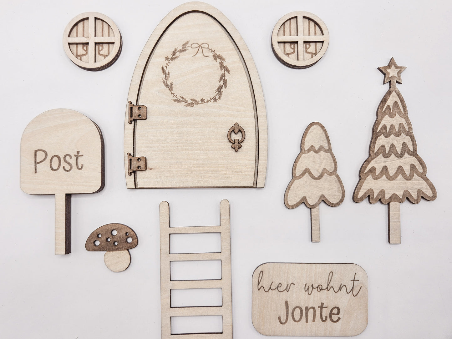 Personalisierbare Holz-Wichteltür mit Leiter, Briefkasten, Pilz und zwei Tannenbäumen an einer weißen Sockelleiste im Kinderzimmer, auf hellblauer Wand mit weißen Punkten montiert.