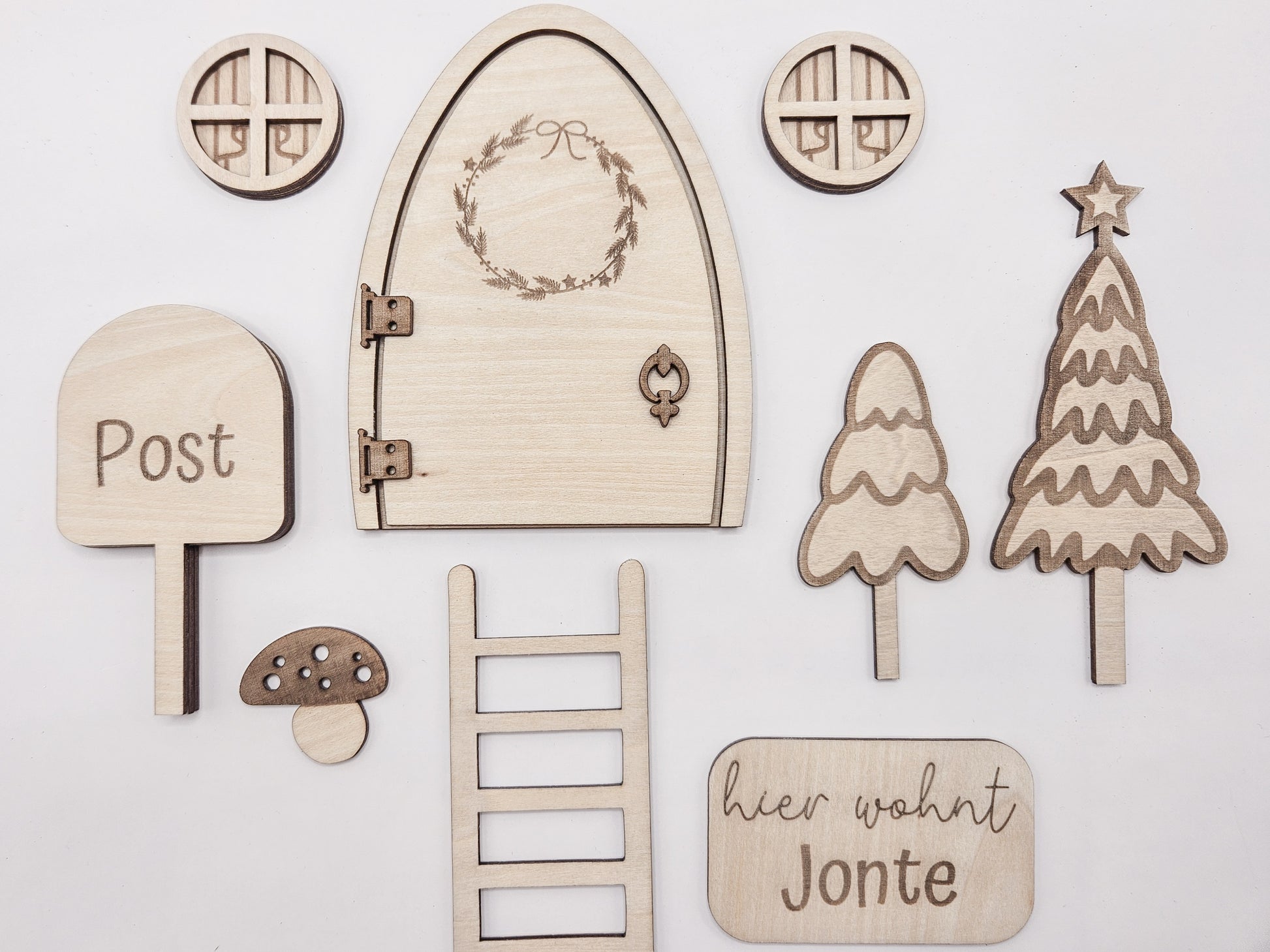 Personalisierbare Holz-Wichteltür mit Leiter, Briefkasten, Pilz und zwei Tannenbäumen an einer weißen Sockelleiste im Kinderzimmer, auf hellblauer Wand mit weißen Punkten montiert.