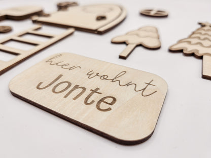 Personalisierbare Holz-Wichteltür mit Leiter, Briefkasten, Pilz und zwei Tannenbäumen an einer weißen Sockelleiste im Kinderzimmer, auf hellblauer Wand mit weißen Punkten montiert.