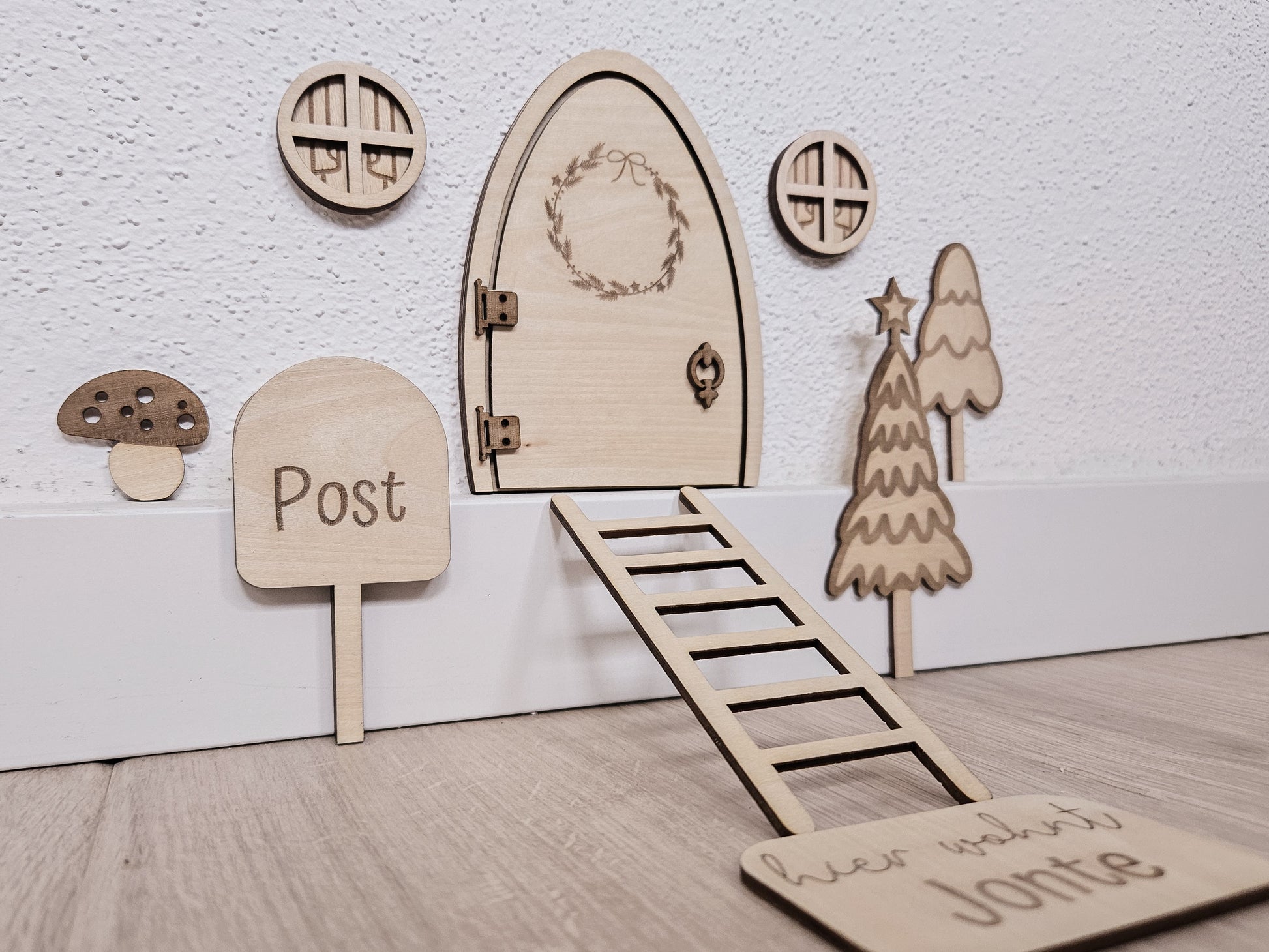 Personalisierbare Holz-Wichteltür mit Leiter, Briefkasten, Pilz und zwei Tannenbäumen an einer weißen Sockelleiste im Kinderzimmer, auf hellblauer Wand mit weißen Punkten montiert.