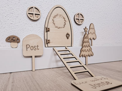 Personalisierbare Holz-Wichteltür mit Leiter, Briefkasten, Pilz und zwei Tannenbäumen an einer weißen Sockelleiste im Kinderzimmer, auf hellblauer Wand mit weißen Punkten montiert.