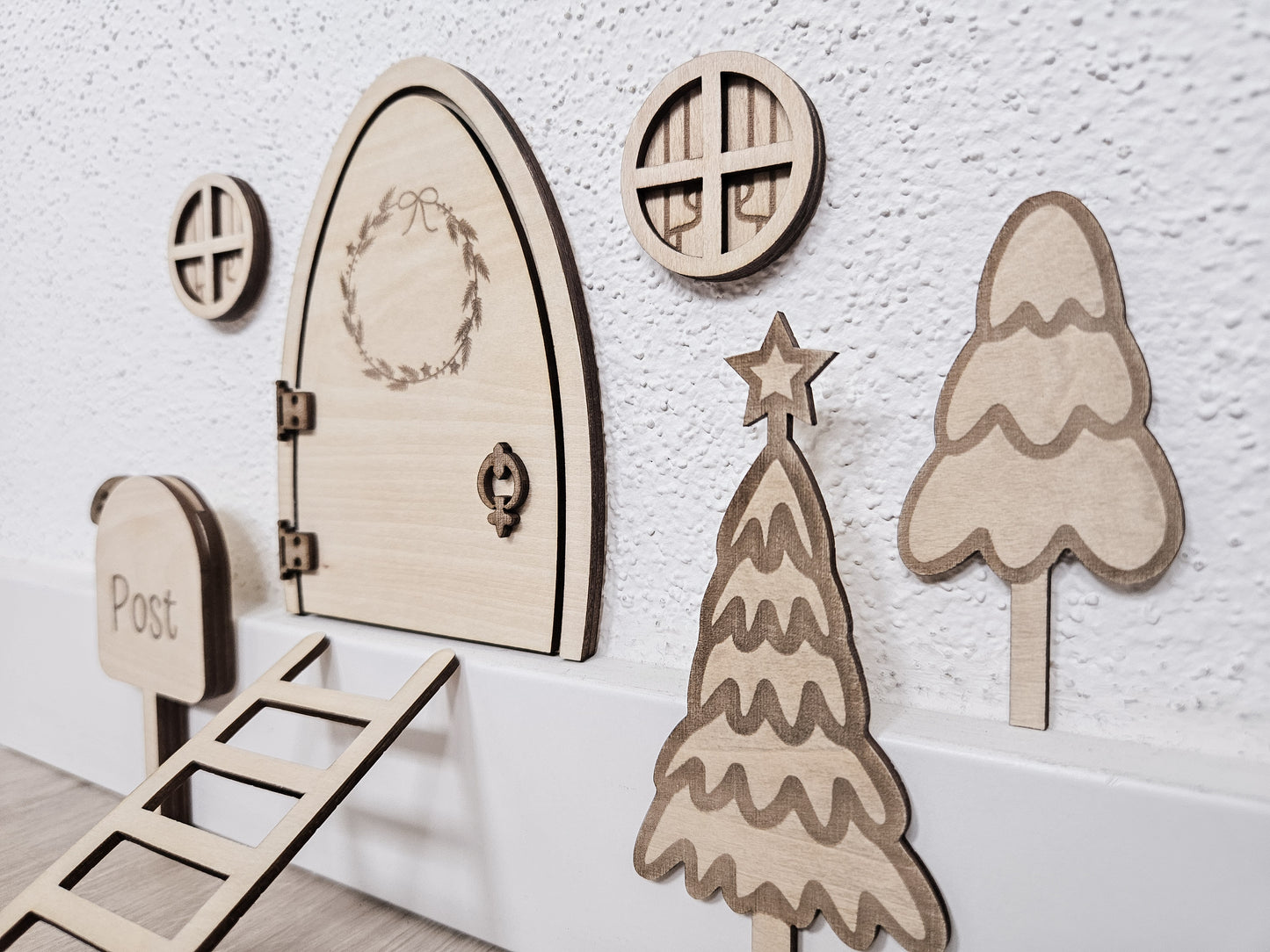 Personalisierbare Holz-Wichteltür mit Leiter, Briefkasten, Pilz und zwei Tannenbäumen an einer weißen Sockelleiste im Kinderzimmer, auf hellblauer Wand mit weißen Punkten montiert.