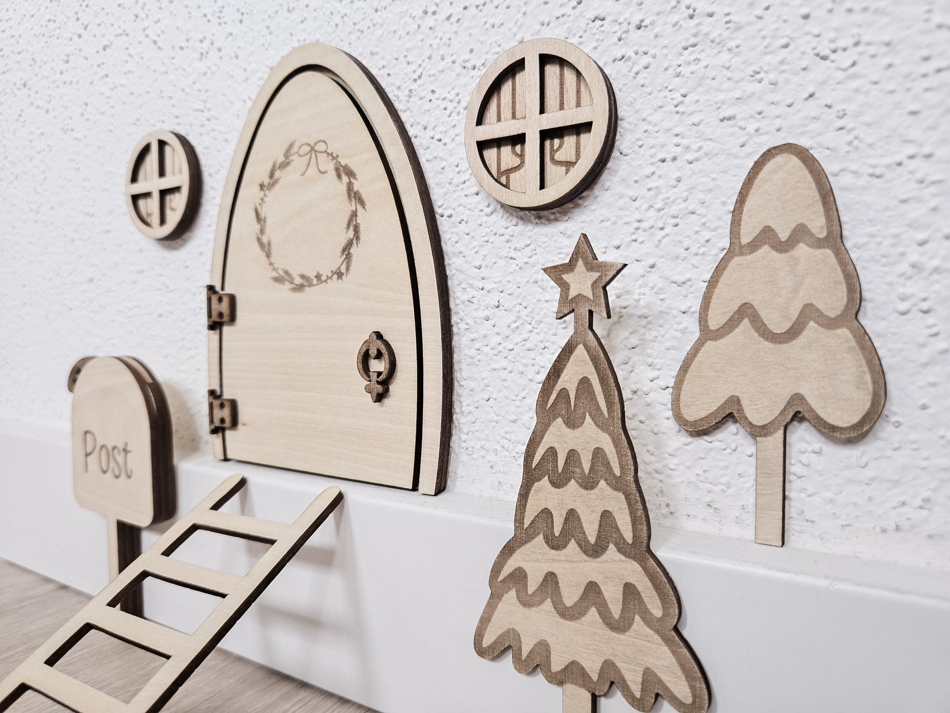 Personalisierbare Holz-Wichteltür mit Leiter, Briefkasten, Pilz und zwei Tannenbäumen an einer weißen Sockelleiste im Kinderzimmer, auf hellblauer Wand mit weißen Punkten montiert.