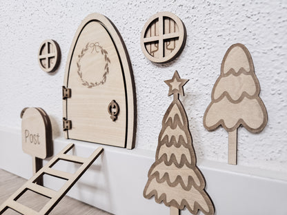 Personalisierbare Holz-Wichteltür mit Leiter, Briefkasten, Pilz und zwei Tannenbäumen an einer weißen Sockelleiste im Kinderzimmer, auf hellblauer Wand mit weißen Punkten montiert.