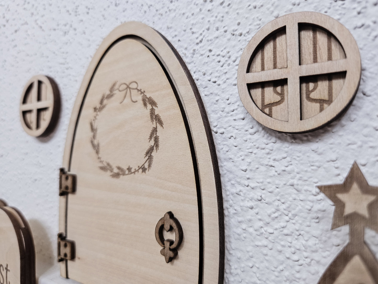 Personalisierbare Holz-Wichteltür mit Leiter, Briefkasten, Pilz und zwei Tannenbäumen an einer weißen Sockelleiste im Kinderzimmer, auf hellblauer Wand mit weißen Punkten montiert.