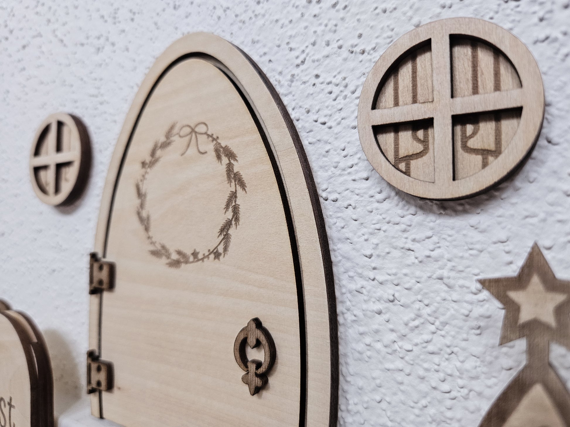 Personalisierbare Holz-Wichteltür mit Leiter, Briefkasten, Pilz und zwei Tannenbäumen an einer weißen Sockelleiste im Kinderzimmer, auf hellblauer Wand mit weißen Punkten montiert.