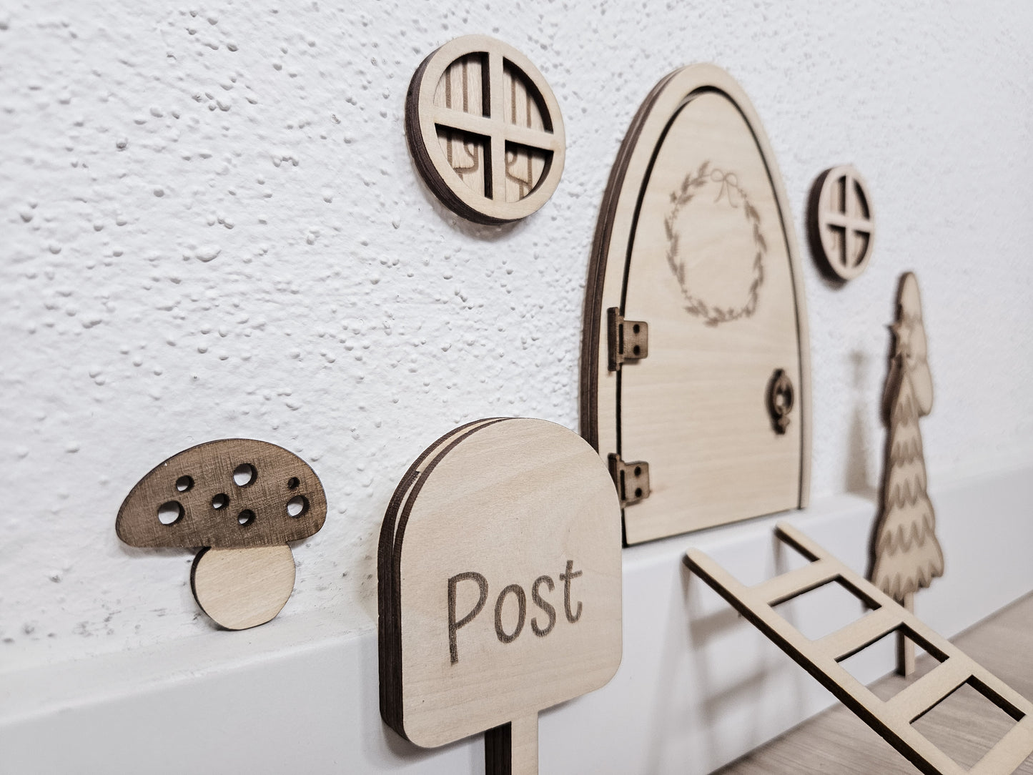 Personalisierbare Holz-Wichteltür mit Leiter, Briefkasten, Pilz und zwei Tannenbäumen an einer weißen Sockelleiste im Kinderzimmer, auf hellblauer Wand mit weißen Punkten montiert.