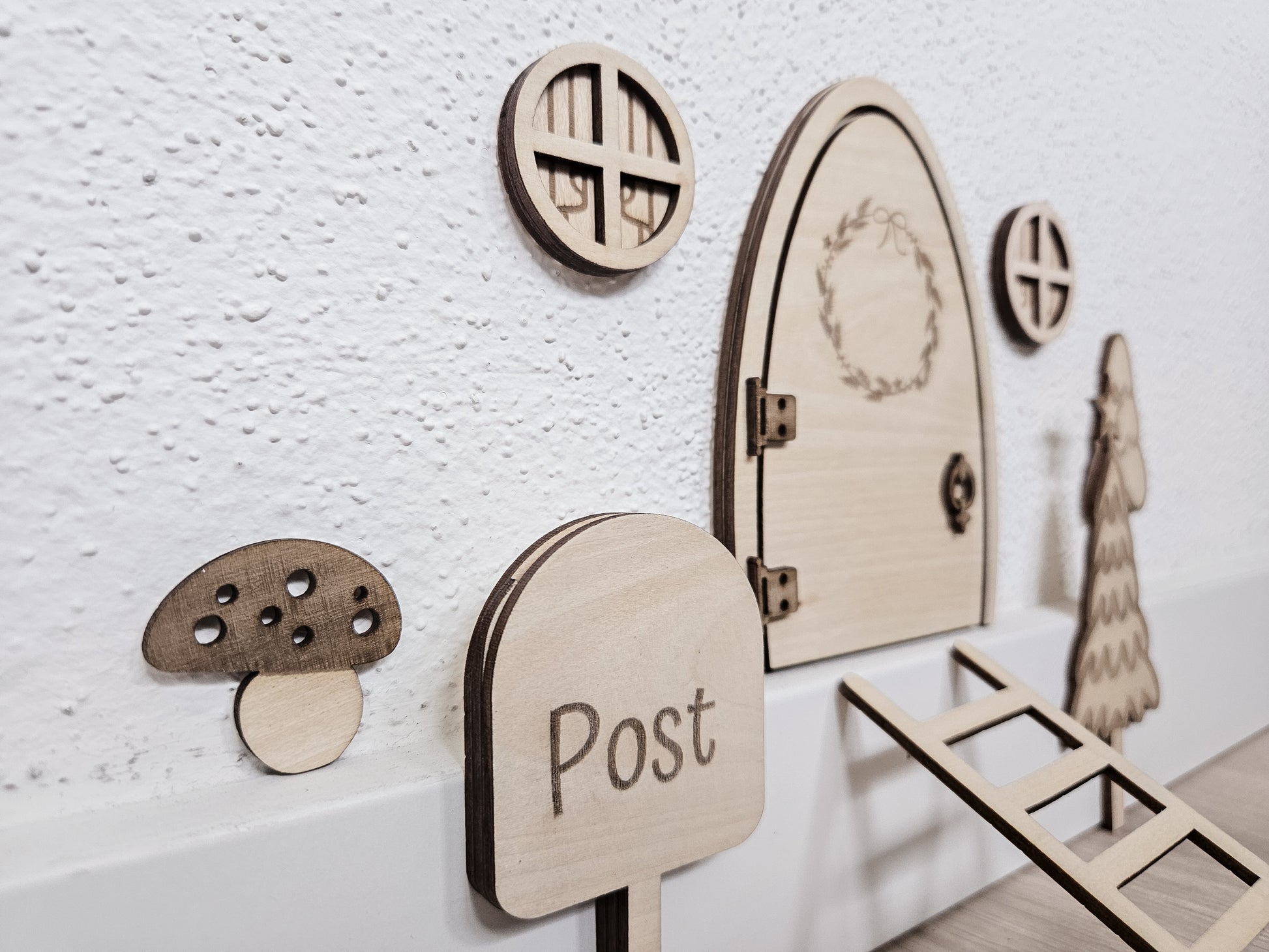 Personalisierbare Holz-Wichteltür mit Leiter, Briefkasten, Pilz und zwei Tannenbäumen an einer weißen Sockelleiste im Kinderzimmer, auf hellblauer Wand mit weißen Punkten montiert.