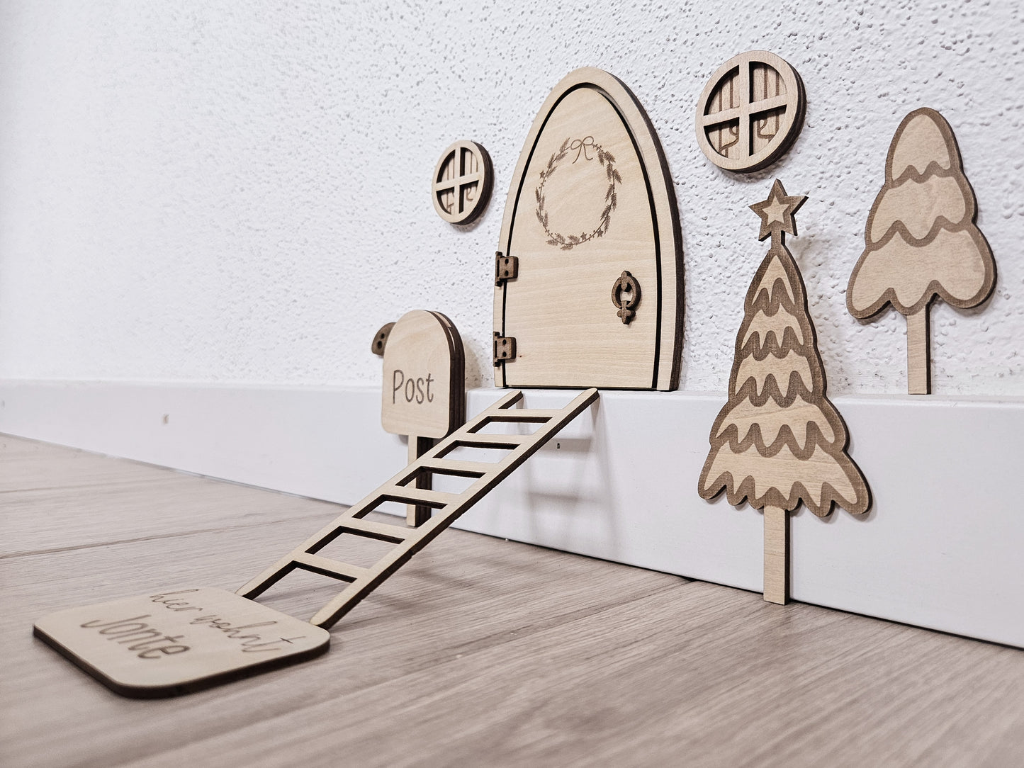 Personalisierbare Holz-Wichteltür mit Leiter, Briefkasten, Pilz und zwei Tannenbäumen an einer weißen Sockelleiste im Kinderzimmer, auf hellblauer Wand mit weißen Punkten montiert.