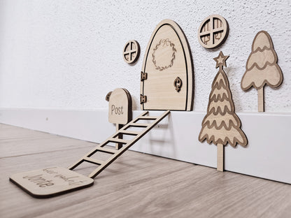 Personalisierbare Holz-Wichteltür mit Leiter, Briefkasten, Pilz und zwei Tannenbäumen an einer weißen Sockelleiste im Kinderzimmer, auf hellblauer Wand mit weißen Punkten montiert.