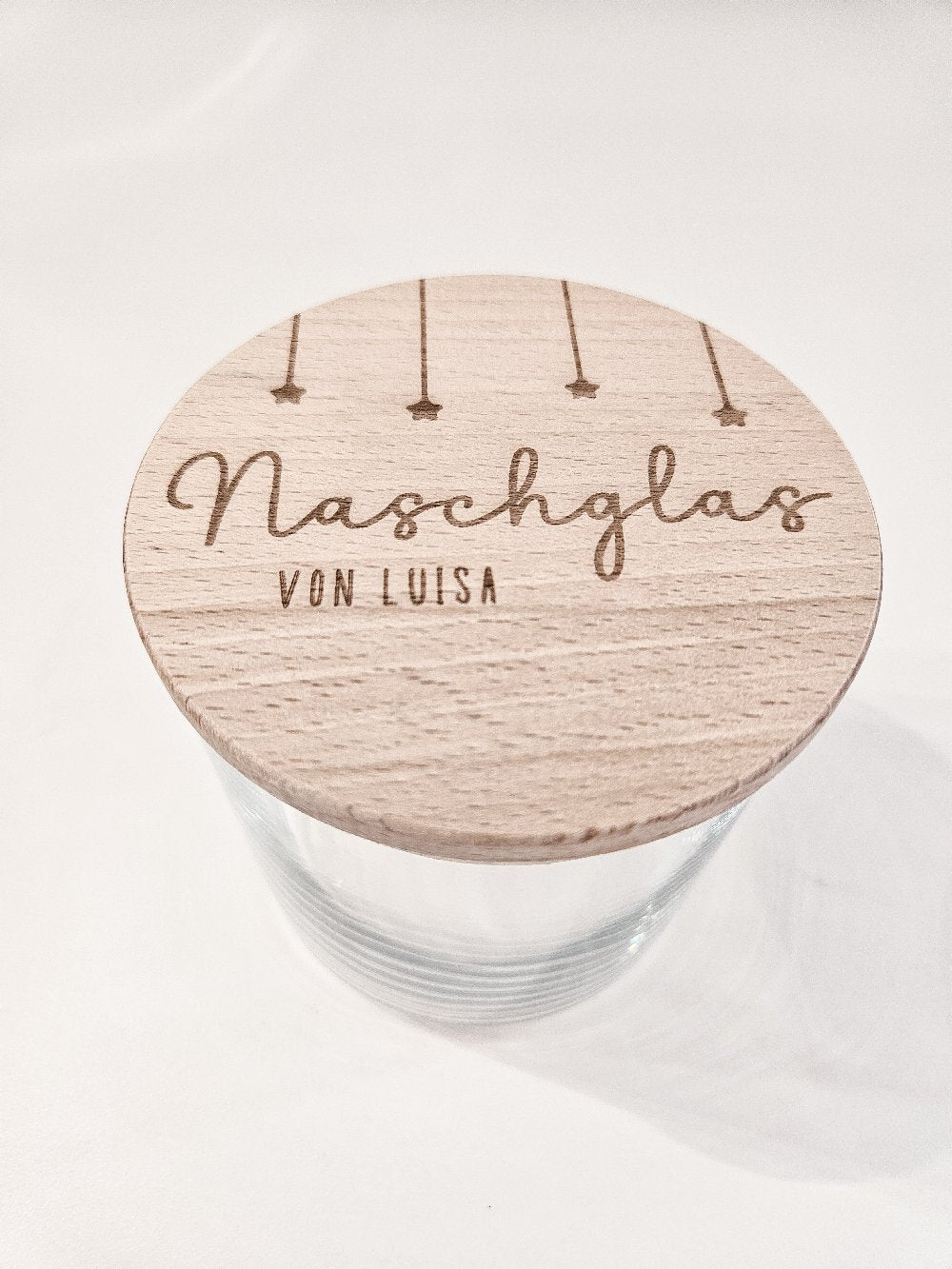 Runder Holzdeckel mit graviertem Schriftzug „Naschglas von Luisa“ und Sternanhängern, auf Glas liegend – personalisierte Geschenkidee.