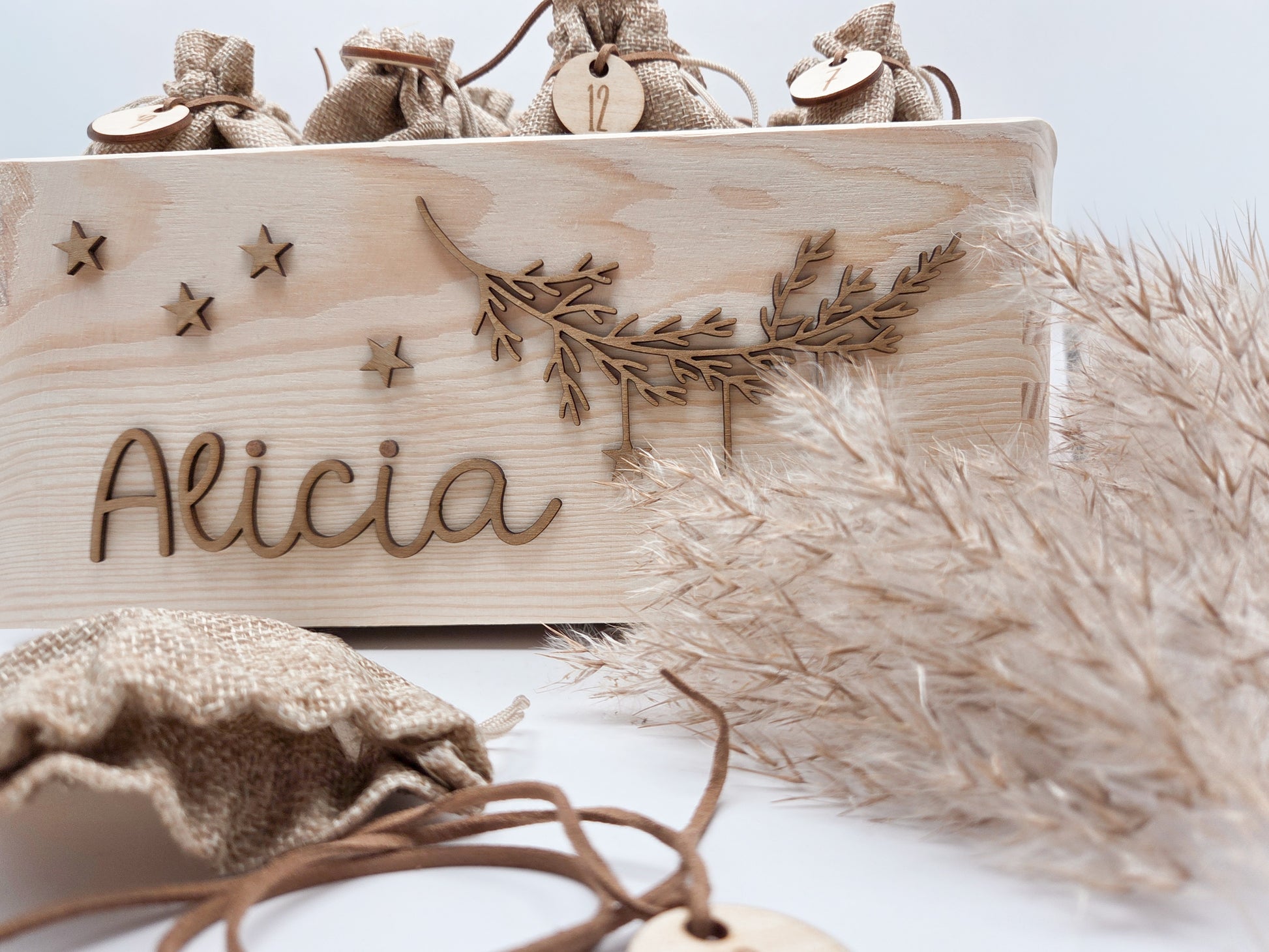 Personalisierte Adventskiste aus hellem Holz mit 3D-Zweig und Sternen, dekoriert mit dem Namen “Alicia”, dazu 24 naturfarbene Jutesäckchen mit gravierten Zahlenanhängern im modernen Scandi-Look.