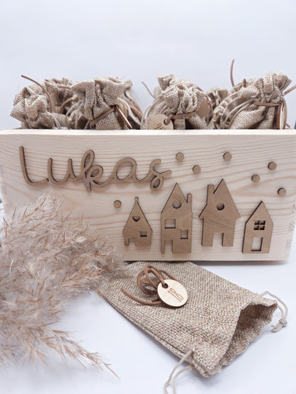 Personalisierte Adventskiste aus Holz mit 3D-Weihnachtsdorf, 24 Jutesäckchen und Zahlenanhängern im hellen skandinavischen Leo&Lu-Stil.