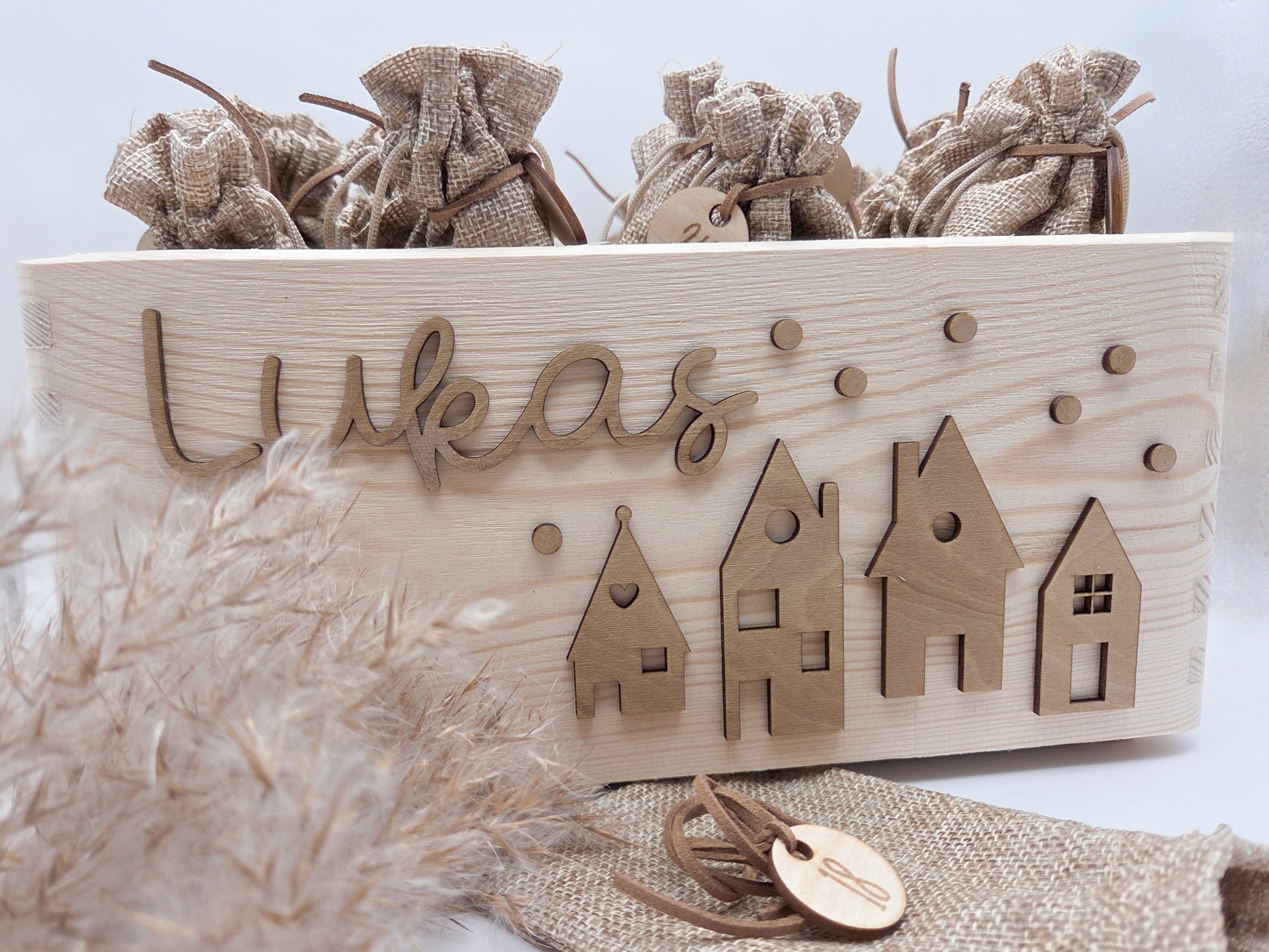 Personalisierte Adventskiste aus Holz mit 3D-Weihnachtsdorf, 24 Jutesäckchen und Zahlenanhängern im hellen skandinavischen Leo&Lu-Stil.