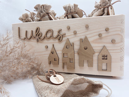 Personalisierte Adventskiste aus Holz mit 3D-Weihnachtsdorf, 24 Jutesäckchen und Zahlenanhängern im hellen skandinavischen Leo&Lu-Stil.