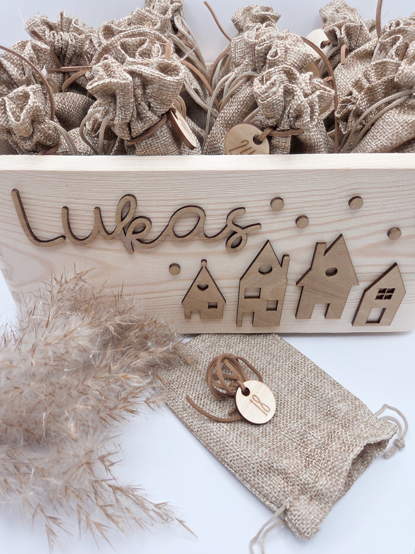 Personalisierte Adventskiste aus Holz mit 3D-Weihnachtsdorf, 24 Jutesäckchen und Zahlenanhängern im hellen skandinavischen Leo&Lu-Stil.
