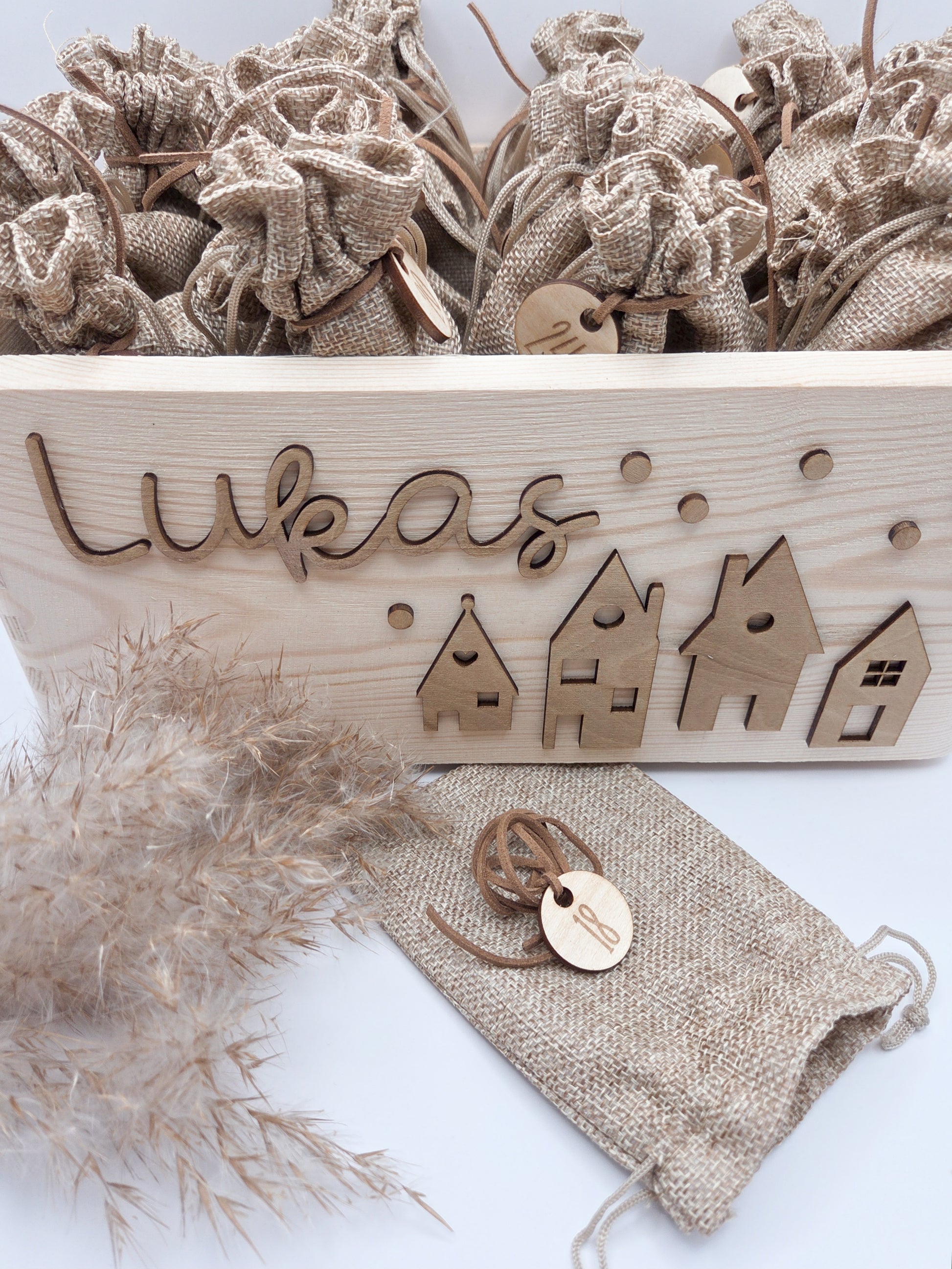 Personalisierte Adventskiste aus Holz mit 3D-Weihnachtsdorf, 24 Jutesäckchen und Zahlenanhängern im hellen skandinavischen Leo&Lu-Stil.