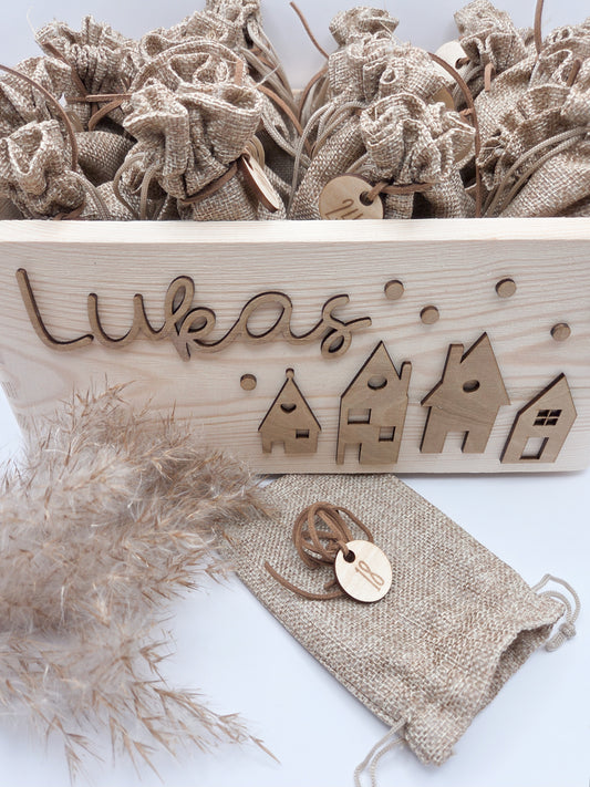 Personalisierte Adventskiste aus Holz mit 3D-Weihnachtsdorf, 24 Jutesäckchen und Zahlenanhängern im hellen skandinavischen Leo&Lu-Stil.
