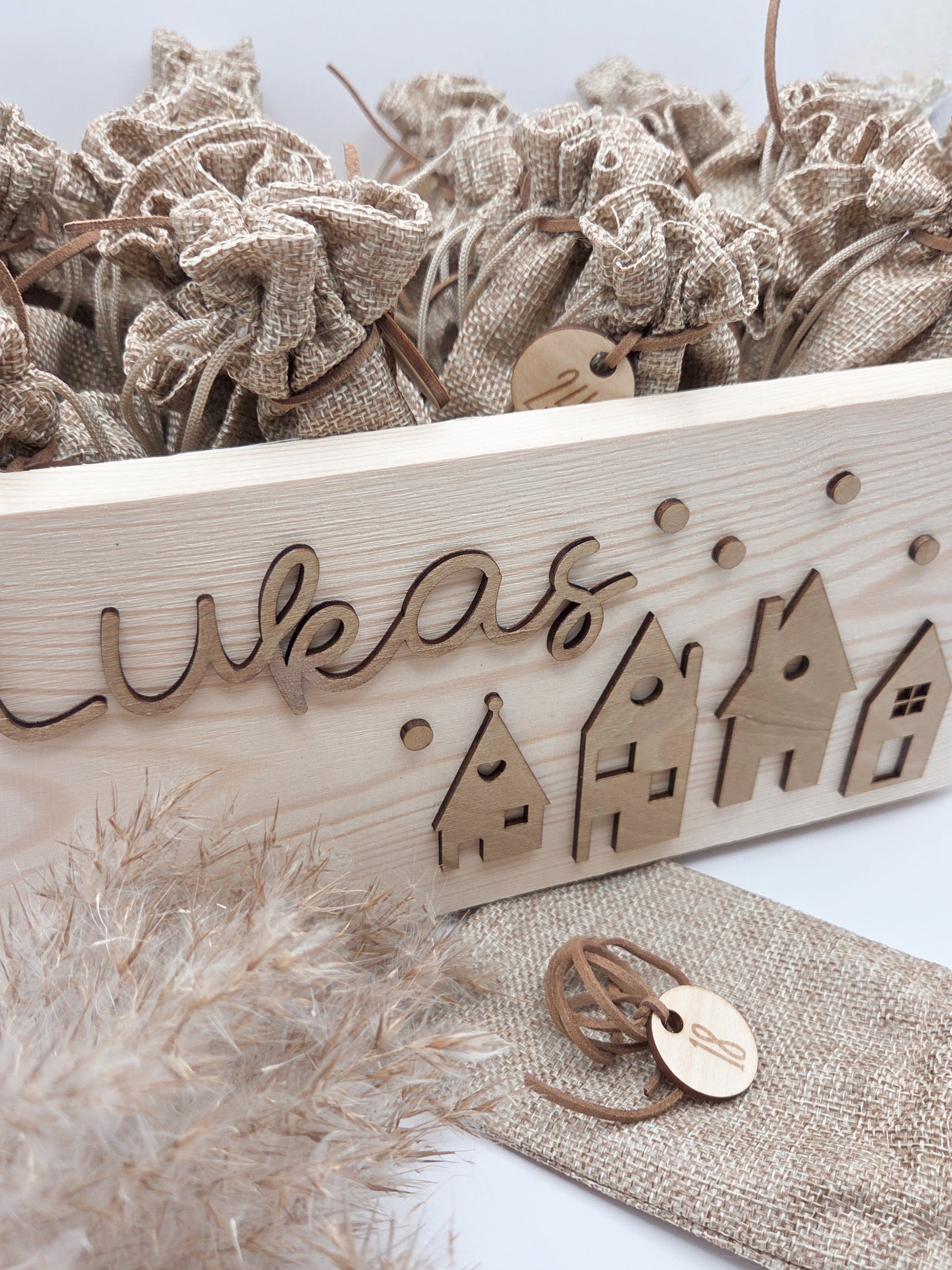 Personalisierte Adventskiste aus Holz mit 3D-Weihnachtsdorf, 24 Jutesäckchen und Zahlenanhängern im hellen skandinavischen Leo&Lu-Stil.