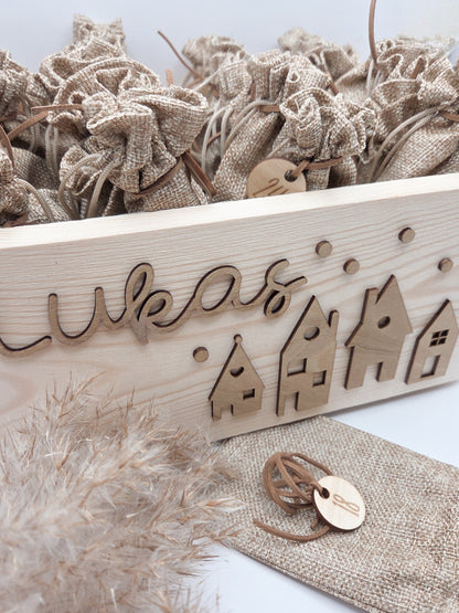 Personalisierte Adventskiste aus Holz mit 3D-Weihnachtsdorf, 24 Jutesäckchen und Zahlenanhängern im hellen skandinavischen Leo&Lu-Stil.