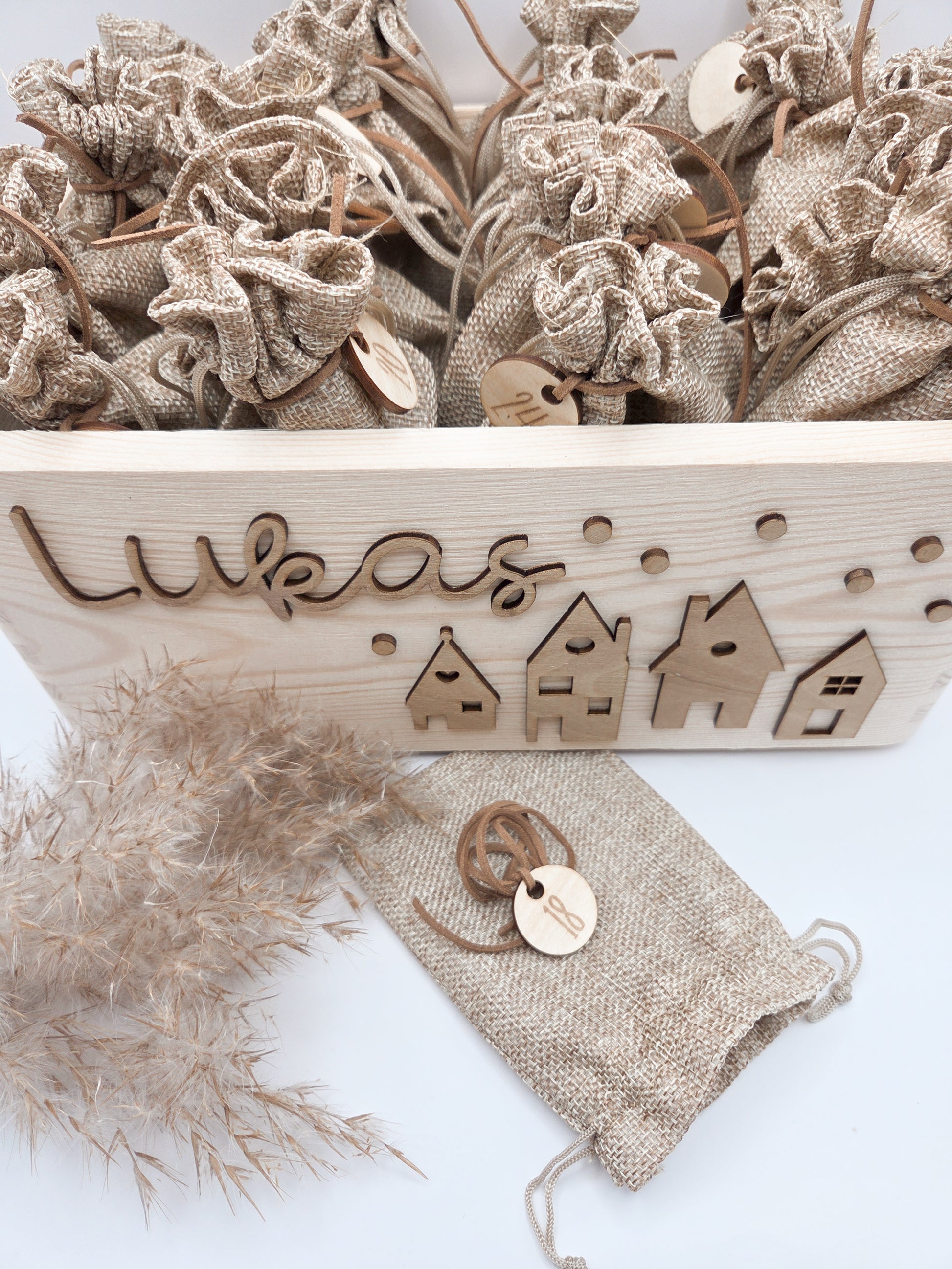 Personalisierte Adventskiste aus Holz mit 3D-Weihnachtsdorf, 24 Jutesäckchen und Zahlenanhängern im hellen skandinavischen Leo&Lu-Stil.