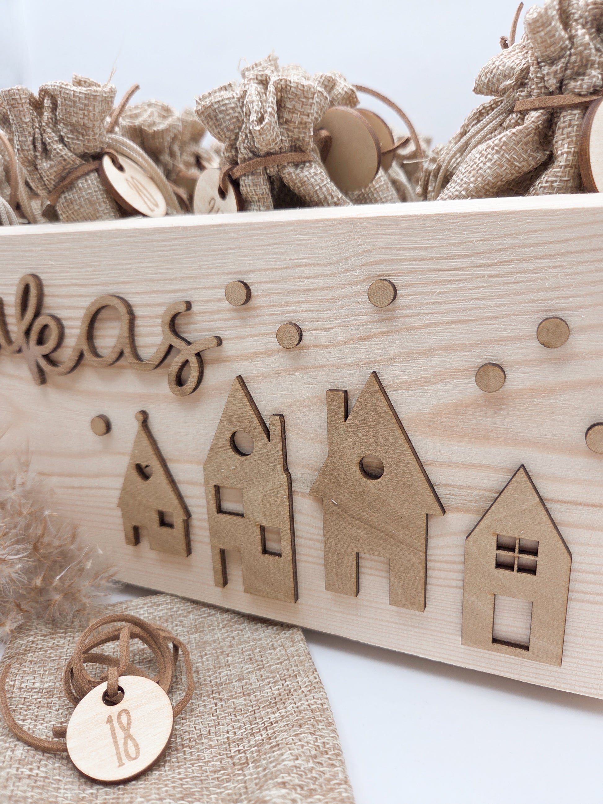 Personalisierte Adventskiste aus Holz mit 3D-Weihnachtsdorf, 24 Jutesäckchen und Zahlenanhängern im hellen skandinavischen Leo&Lu-Stil.