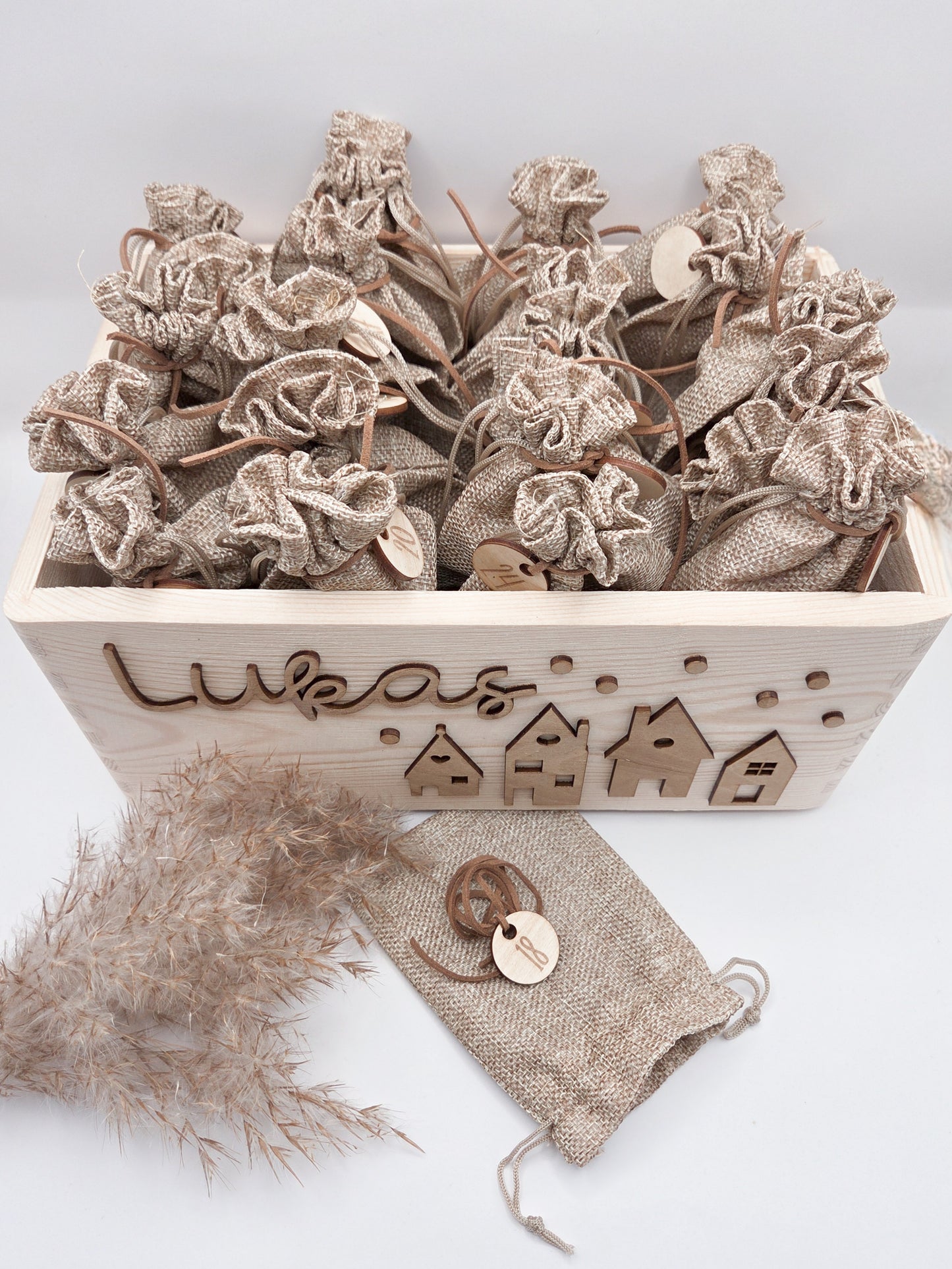 Personalisierte Adventskiste aus Holz mit 3D-Weihnachtsdorf, 24 Jutesäckchen und Zahlenanhängern im hellen skandinavischen Leo&Lu-Stil.