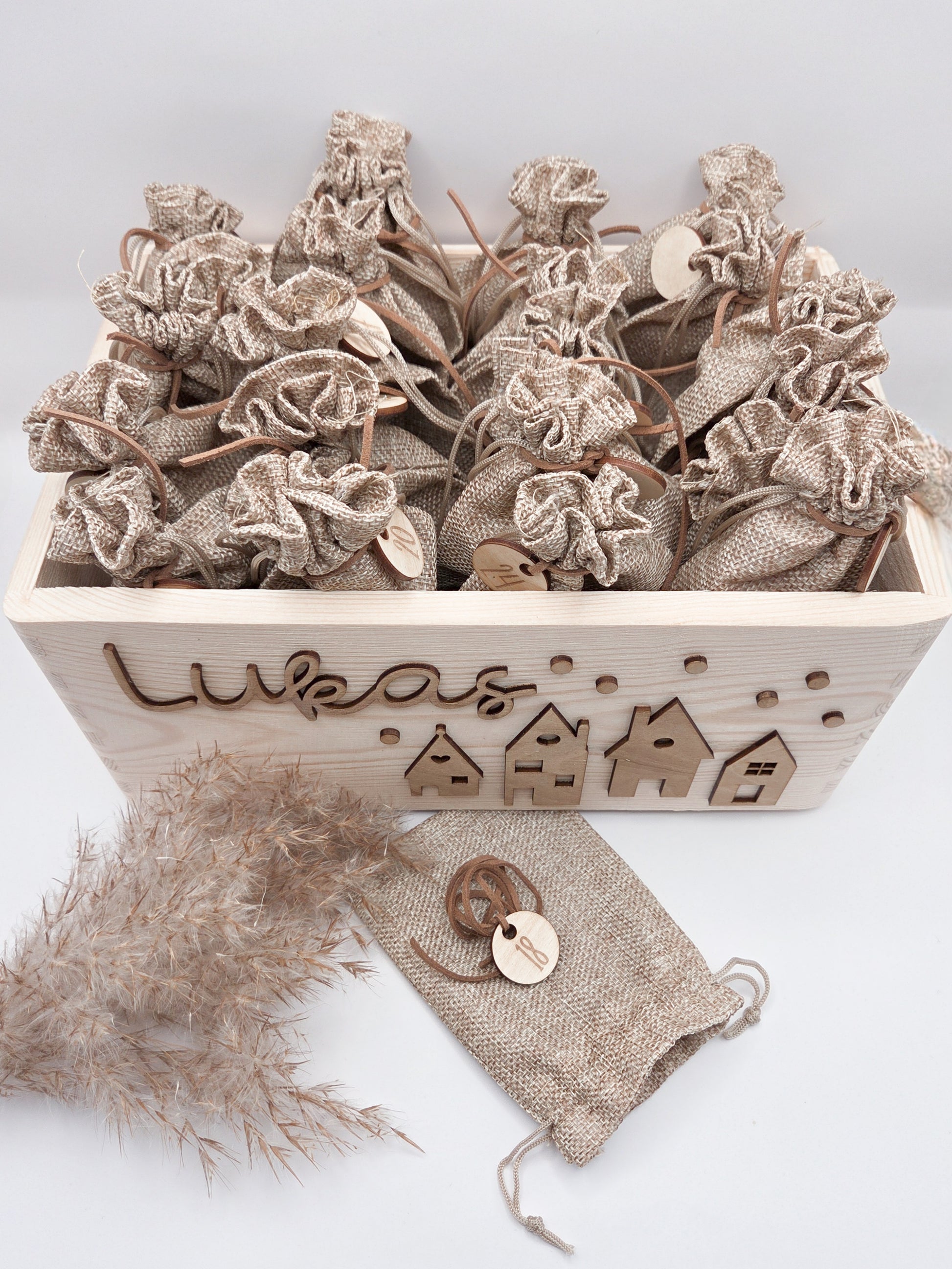 Personalisierte Adventskiste aus Holz mit 3D-Weihnachtsdorf, 24 Jutesäckchen und Zahlenanhängern im hellen skandinavischen Leo&Lu-Stil.