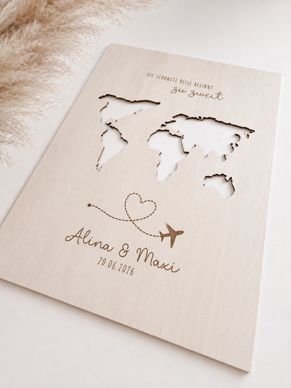 Personalisiertes Geldgeschenk zur Hochzeit aus hellem Holz mit filigraner Weltkarte und eingesteckten Euro-Geldscheinen hinter den Kontinenten. Die gravierte Aufschrift „Die schönste Reise beginnt zu zweit“ sowie die Namen des Brautpaares und das Hochzeitsdatum machen dieses Hochzeitsgeschenk zu einer besonderen Erinnerung. Das Holzbild steht in einer romantischen Hochzeitskulisse mit weißen Rosen, Kerzenlicht, Eheringen und eleganter Tischdekoration. Perfekt als Geldgeschenk für Flitterwochen, Reisekasse o