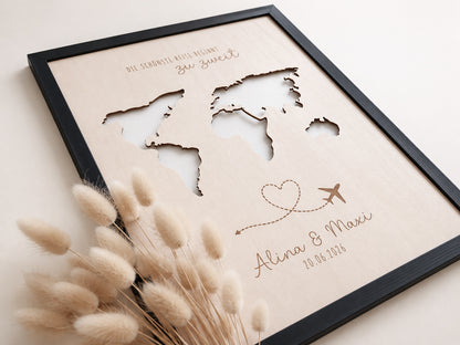 Personalisiertes Geldgeschenk zur Hochzeit aus hellem Holz mit filigraner Weltkarte und eingesteckten Euro-Geldscheinen hinter den Kontinenten. Die gravierte Aufschrift „Die schönste Reise beginnt zu zweit“ sowie die Namen des Brautpaares und das Hochzeitsdatum machen dieses Hochzeitsgeschenk zu einer besonderen Erinnerung. Das Holzbild steht in einer romantischen Hochzeitskulisse mit weißen Rosen, Kerzenlicht, Eheringen und eleganter Tischdekoration. Perfekt als Geldgeschenk für Flitterwochen, Reisekasse o