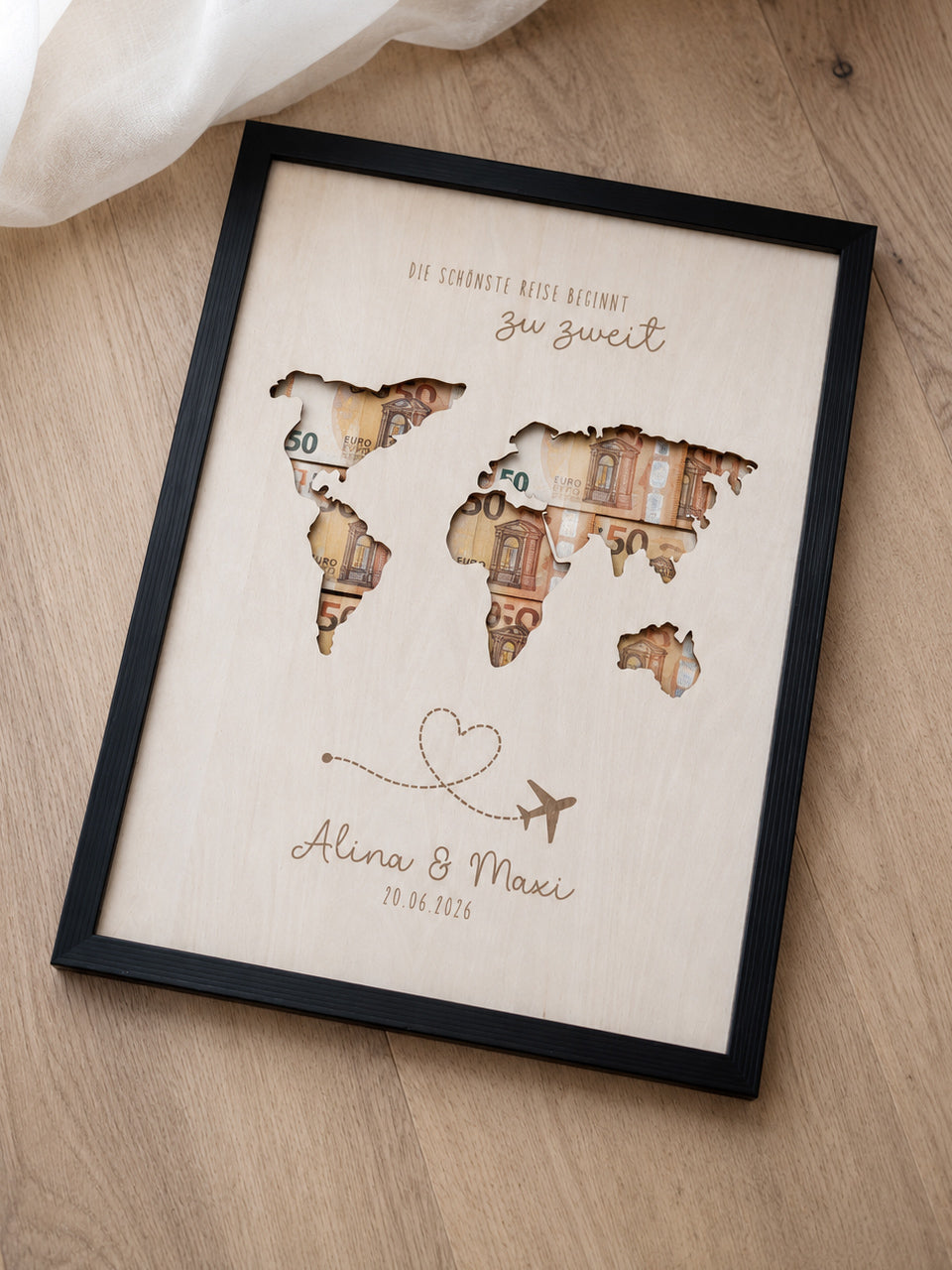 Personalisiertes Geldgeschenk zur Hochzeit aus hellem Holz mit filigraner Weltkarte und eingesteckten Euro-Geldscheinen hinter den Kontinenten. Die gravierte Aufschrift „Die schönste Reise beginnt zu zweit“ sowie die Namen des Brautpaares und das Hochzeitsdatum machen dieses Hochzeitsgeschenk zu einer besonderen Erinnerung. Das Holzbild steht in einer romantischen Hochzeitskulisse mit weißen Rosen, Kerzenlicht, Eheringen und eleganter Tischdekoration. Perfekt als Geldgeschenk für Flitterwochen, Reisekasse o