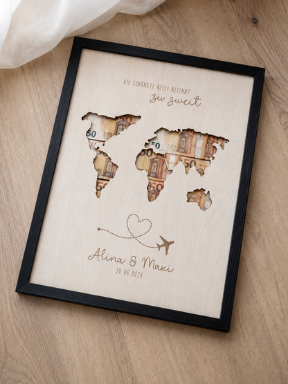 Personalisiertes Geldgeschenk zur Hochzeit aus hellem Holz mit filigraner Weltkarte und eingesteckten Euro-Geldscheinen hinter den Kontinenten. Die gravierte Aufschrift „Die schönste Reise beginnt zu zweit“ sowie die Namen des Brautpaares und das Hochzeitsdatum machen dieses Hochzeitsgeschenk zu einer besonderen Erinnerung. Das Holzbild steht in einer romantischen Hochzeitskulisse mit weißen Rosen, Kerzenlicht, Eheringen und eleganter Tischdekoration. Perfekt als Geldgeschenk für Flitterwochen, Reisekasse o