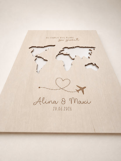Personalisiertes Geldgeschenk zur Hochzeit aus hellem Holz mit filigraner Weltkarte und eingesteckten Euro-Geldscheinen hinter den Kontinenten. Die gravierte Aufschrift „Die schönste Reise beginnt zu zweit“ sowie die Namen des Brautpaares und das Hochzeitsdatum machen dieses Hochzeitsgeschenk zu einer besonderen Erinnerung. Das Holzbild steht in einer romantischen Hochzeitskulisse mit weißen Rosen, Kerzenlicht, Eheringen und eleganter Tischdekoration. Perfekt als Geldgeschenk für Flitterwochen, Reisekasse o